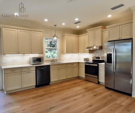 A freshly remodeled classy modern kitchen featuring oak floors, stainless steel appliances, and cream colored cabinets. Warm recessed LED lighting and a picture window over the sink light up the space.