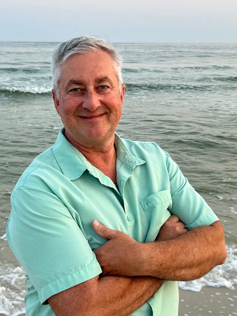 The owner of Green Coast Living, smiles while facing the viewer. The calm surf of Gulf shores beach rolls in the background.